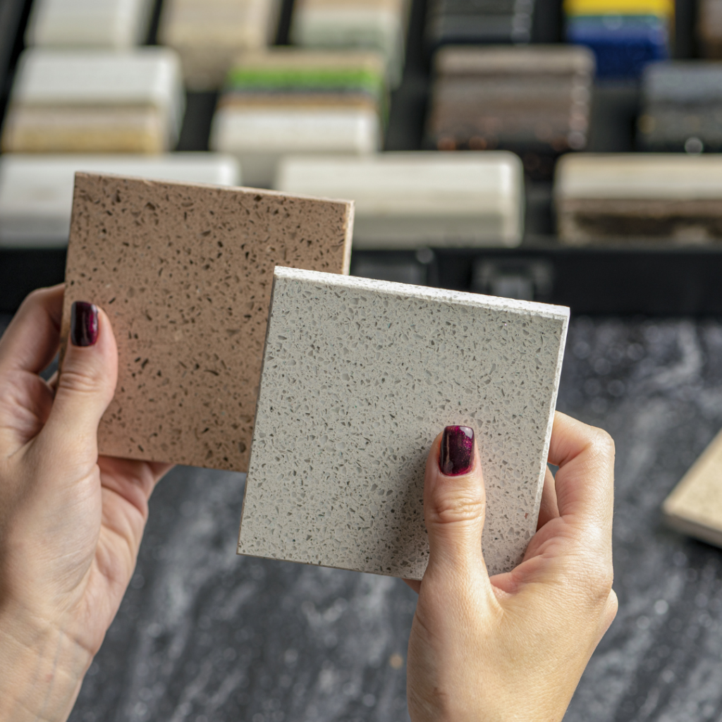 Cost quartz: Hands holding two quartz countertop samples in beige and light gray with a display of additional samples in the background.