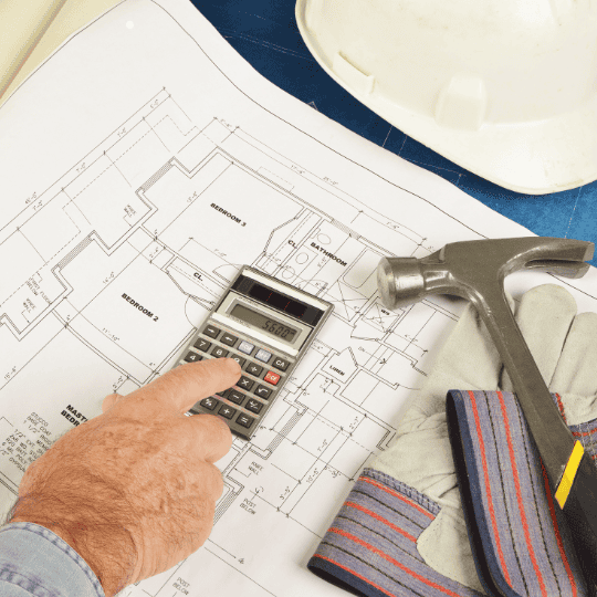 Worker using a calculator to estimate condo kitchen renovation cost with floor plans, a hammer, gloves, and a hard hat on the table.